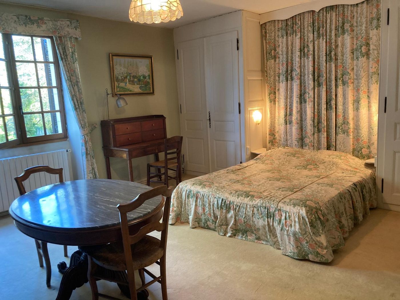 Chambre Marguerite : grandes fenêtres sur les arbres, table ronde et lit à volants fleuris