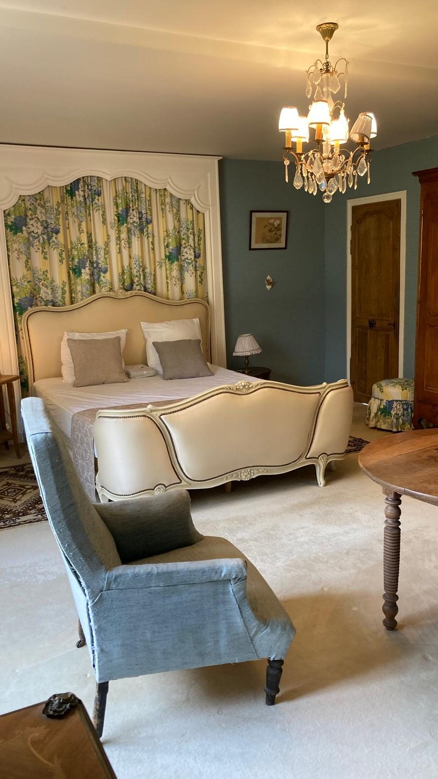 Chambre Jacynthe : lustre à pampilles, lit drapé de tissu fleuri et fauteuil bleu-gris